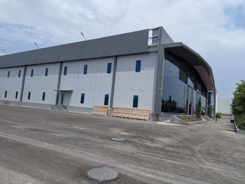 Modern industrial building with white and dark gray metal cladding, blue window panels, and a paved parking area on a partly cloudy day