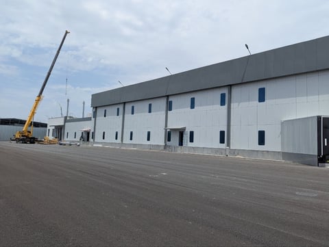 Industrial warehouse under construction with white metal paneling, blue windows, and yellow crane equipment on asphalt lot