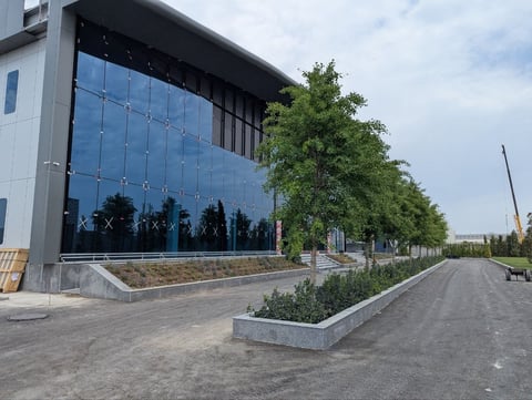 Modern building with large blue-tinted glass facade and concrete planter boxes along a tree-lined paved pathway.